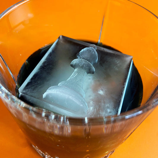 A clear glass with a black ice mold designed to cool drinks, shaped like a chess piece, placed on an orange surface.