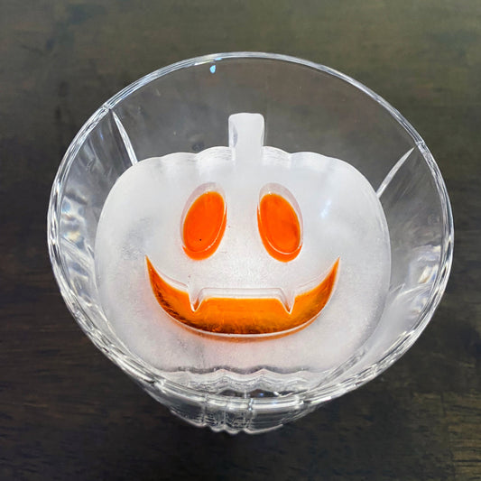 An ice cube mold in the shape of a smiling pumpkin with orange and white colors, placed inside a clear cocktail glass.