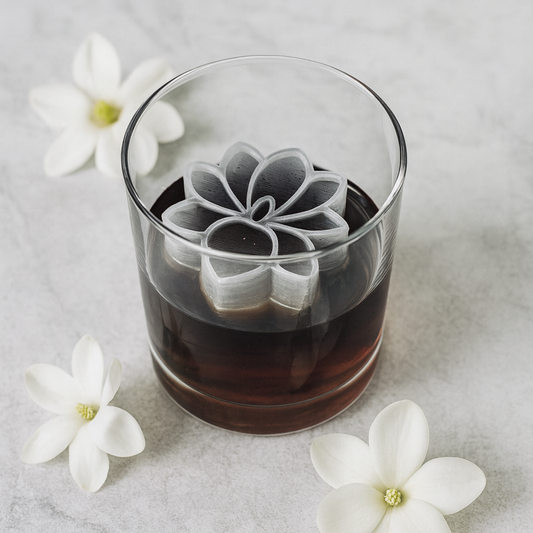 Glass of dark liquid with a magnolia flower-shaped ice cube, surrounded by white flowers on a light gray background