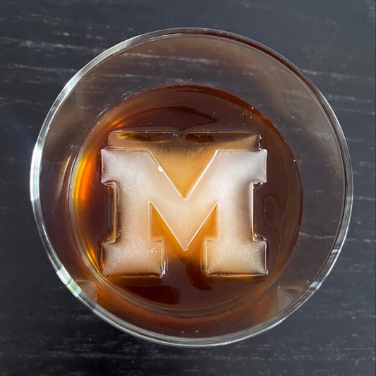 An ice cube with the letter 'M' inscribed, floating in a glass of whiskey.