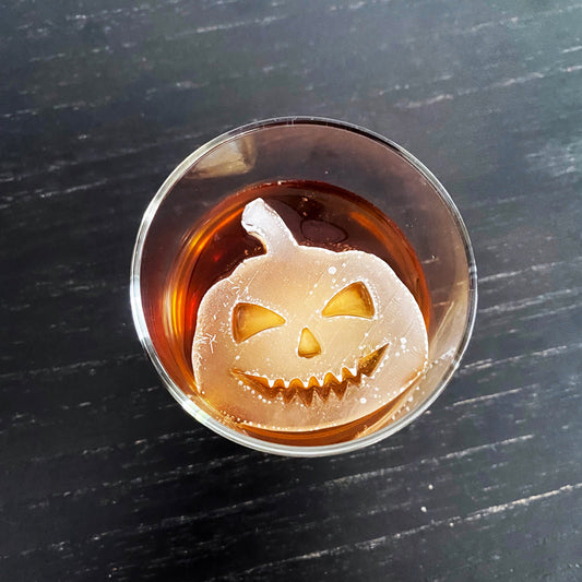 A pumpkin-shaped ice mold in a glass filled with a brown liquid, likely to be used for creating Halloween-themed ice cubes.
