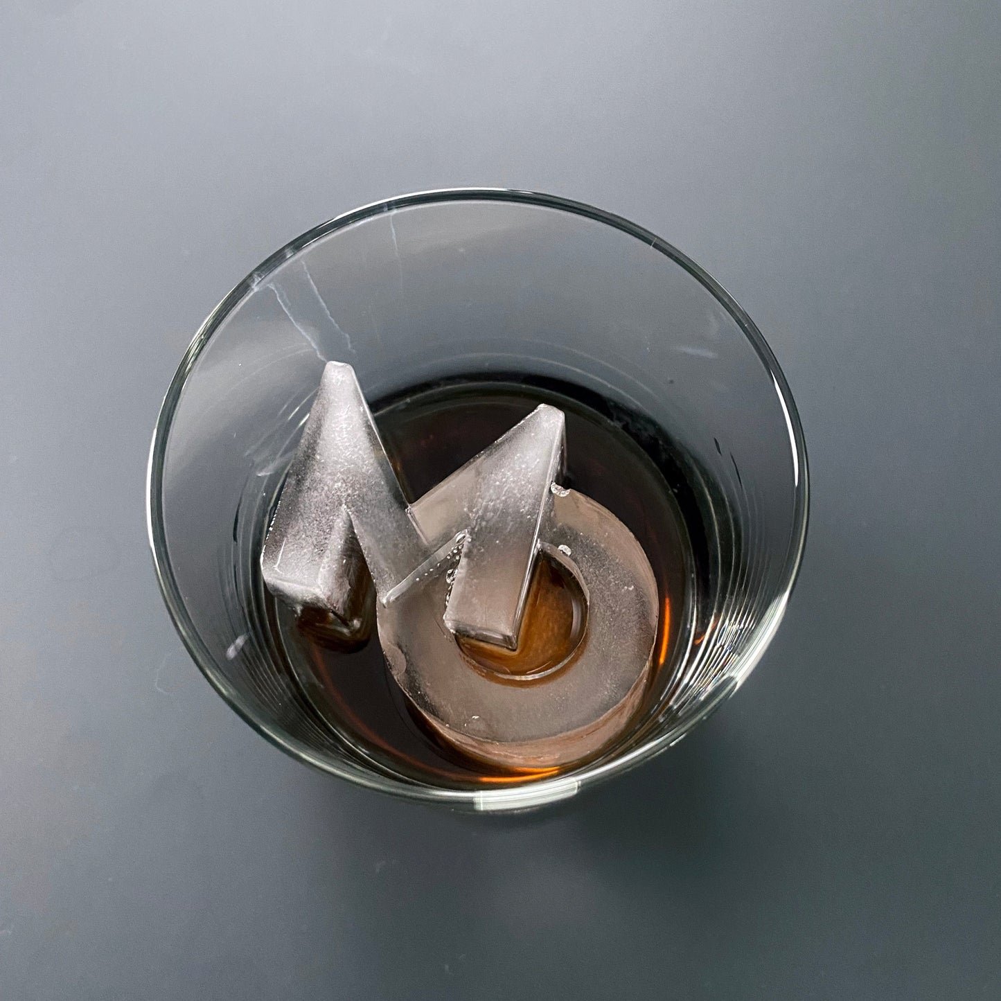 A clear glass with a letter 'M' ice cube inside, on a grey background.