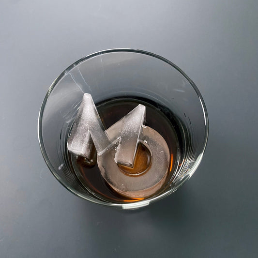 A clear glass with a letter 'M' ice cube inside, on a grey background.