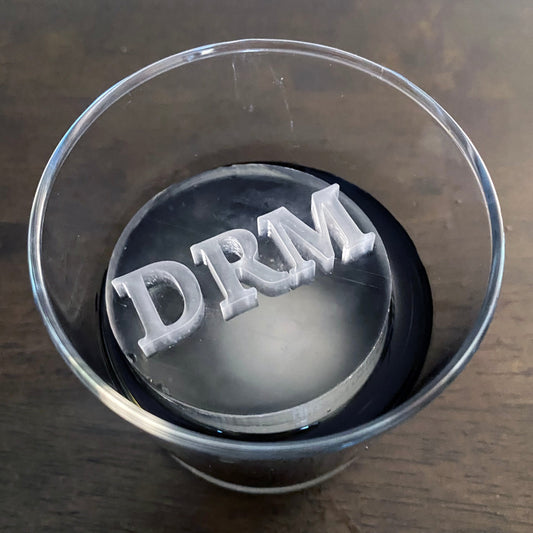 Clear drinks glass with a cylinder ice cube featuring the letters 'DRM' on a wooden surface.