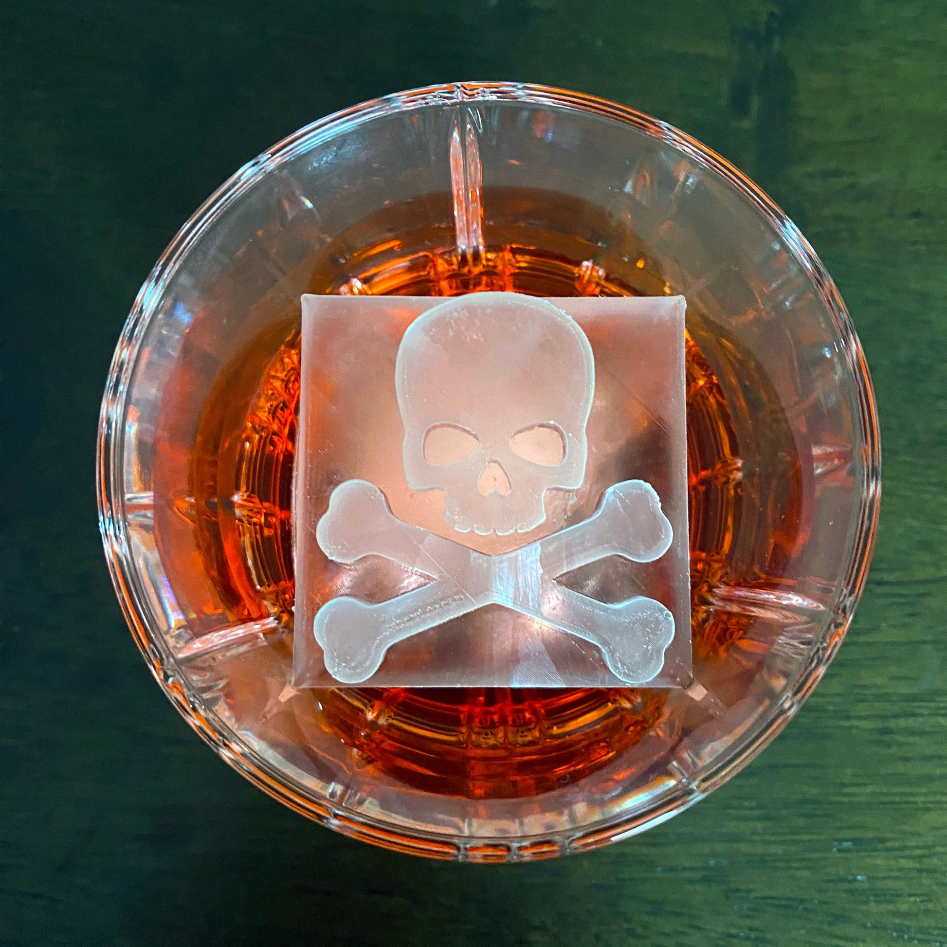 Clear glass with a skull and crossbones design partially submerged in amber liquid on a dark green background